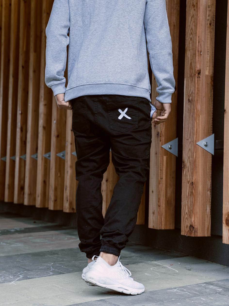 Mężczyzna ubrany w czarne joggery w stylu streetwear od marki Point X Pocket. Spodnie są materiałowe ze ściągaczem przy kostce i białym logo X na tylnej kieszeni